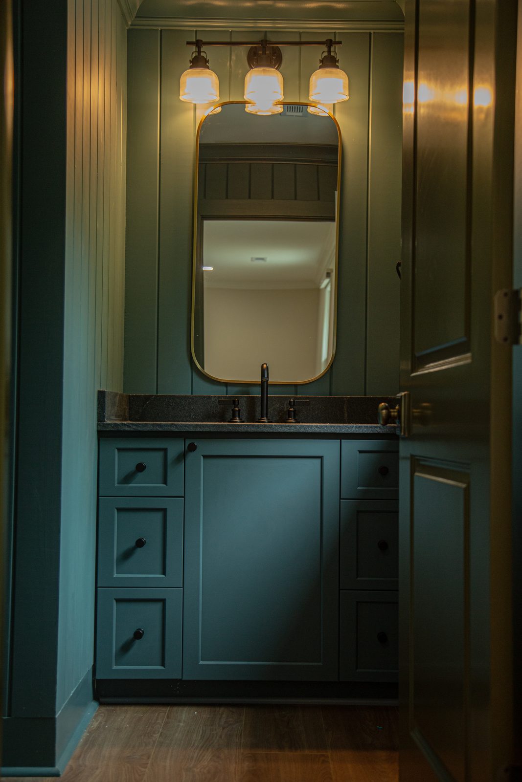 Custom powder room with deep green paneling, a brass mirror, and a stone vanity top